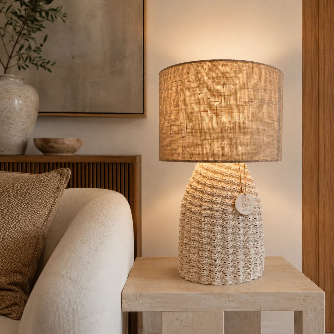 Lampada da tavolo Fira accesa con paralume in juta naturale e base intrecciata, posizionata accanto a un divano in ambiente moderno.