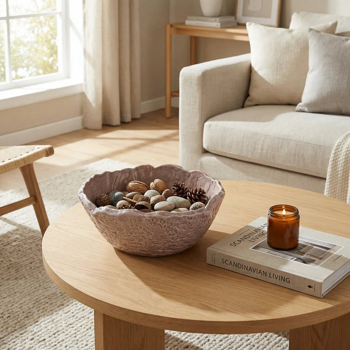 Ciotola in ceramica artigianale con pigne e sassi su tavolino in legno accanto a candela e libro in soggiorno luminoso