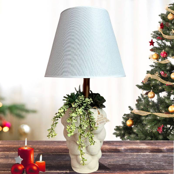 Lampada decorativa con base a forma di volto, piante verdi e paralume chiaro su tavolo natalizio.