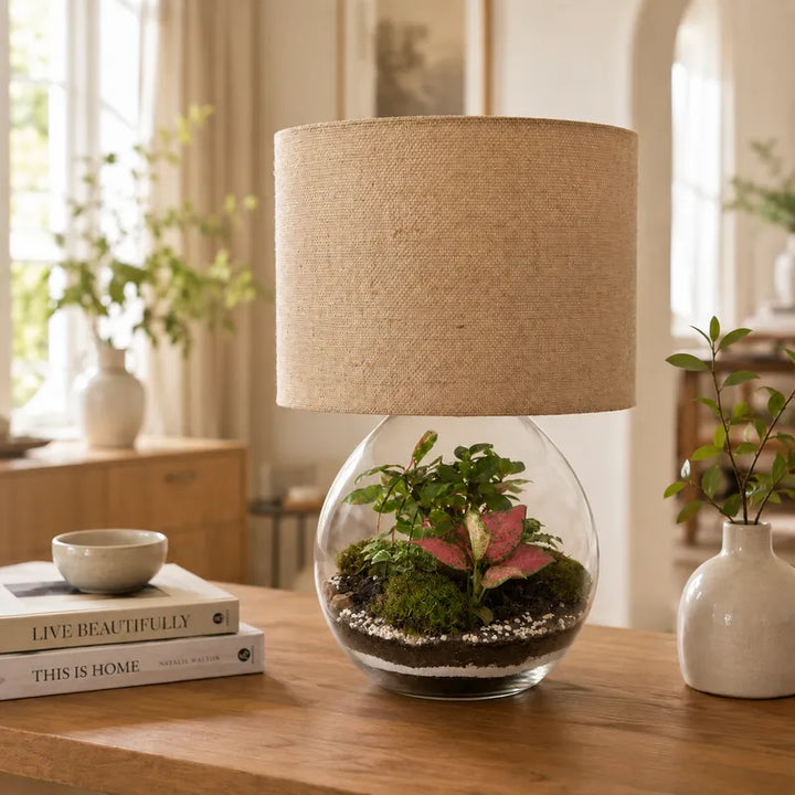 Lamparium Aida con paralume cilindrico in juta e base terrarium in vetro con piante verdi, Fittonia rosa e muschio, inserito in un soggiorno luminoso.
