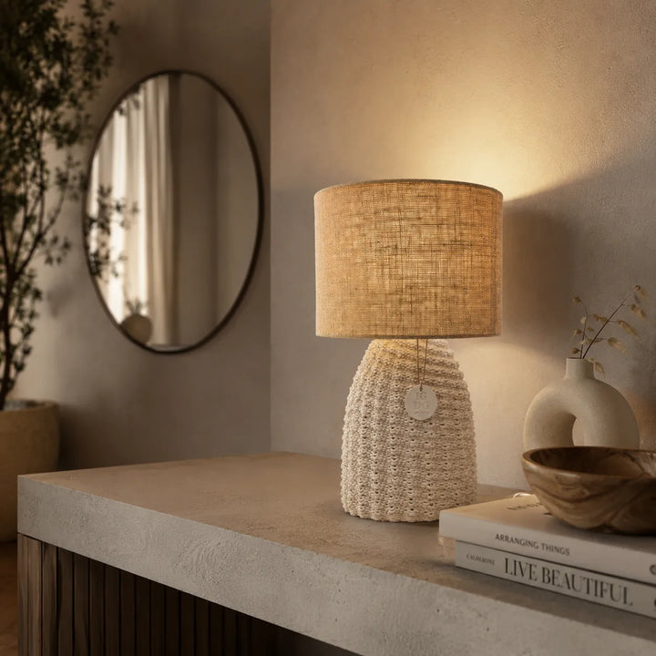 Lampada da tavolo Fira con base intrecciata color avorio e paralume cilindrico in juta naturale su sfondo neutro.
