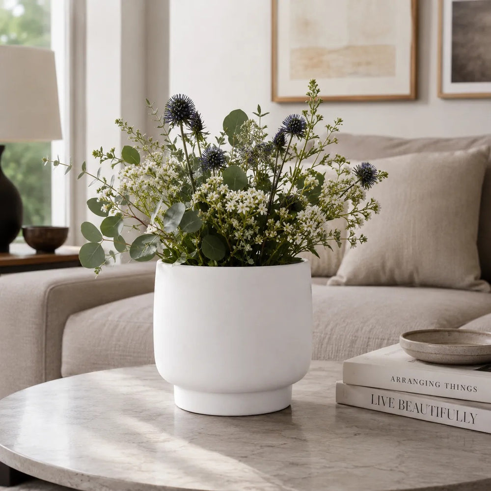 Vaso Sweet bianco dal design minimalista con composizione di fiori bianchi, cardo blu ed eucalipto, posizionato su tavolino da salotto moderno.