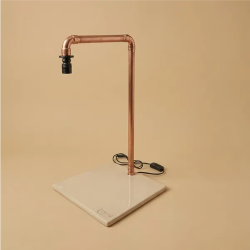 Lampada LED da tavolo industriale in tubo di rame con base quadrata in ceramica artigianale color crema, interruttore a filo nero su sfondo beige
