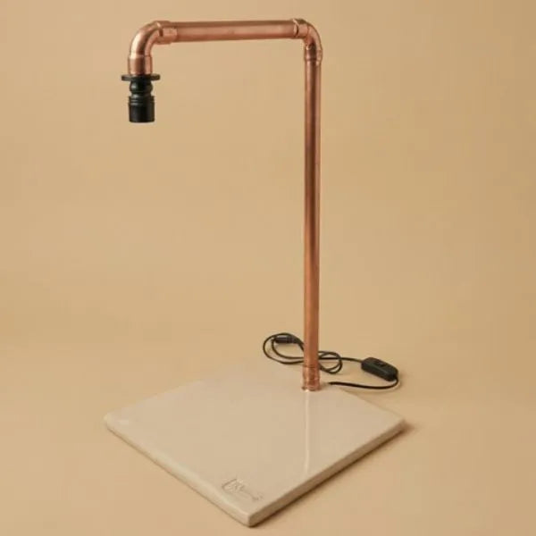 Lampada LED da tavolo industriale in tubo di rame con base quadrata in ceramica artigianale color crema, interruttore a filo nero su sfondo beige
